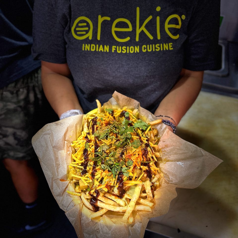 Person wearing an Arekie Indian Fusion Cuisine shirt holds loaded fries topped with chutneys, shredded cheese, spices, and cilantro.